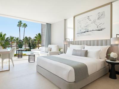 Helles Hotelzimmer mit großem Bett, Sitzbereich und Blick auf Pool und Palmen.
