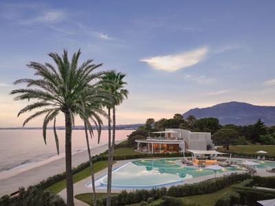 Strandhotel mit Palmen, Pool und Bergblick bei Sonnenuntergang.