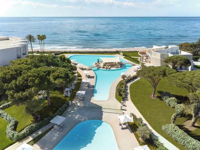 Hotel mit zwei Swimmingpools, grüner Gartenanlage und Blick auf das Meer im Hintergrund.