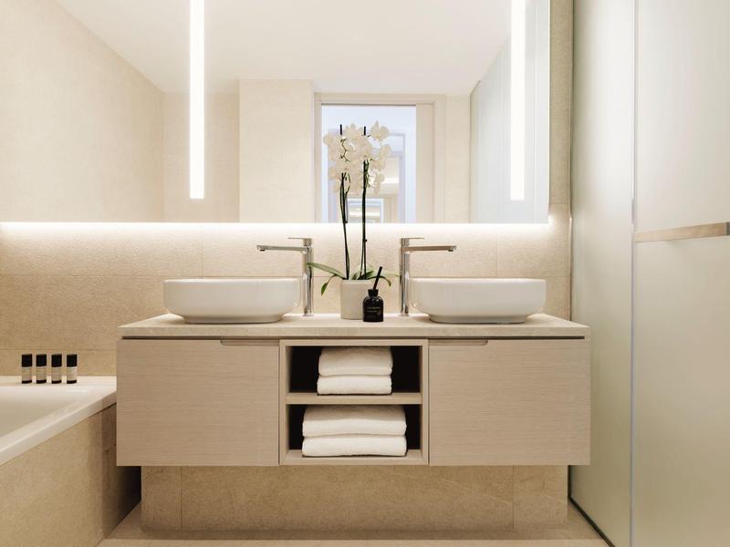 Modernes Badezimmer mit Doppelwaschbecken, beleuchtetem Spiegel und hellen cremefarbenen Fliesen.