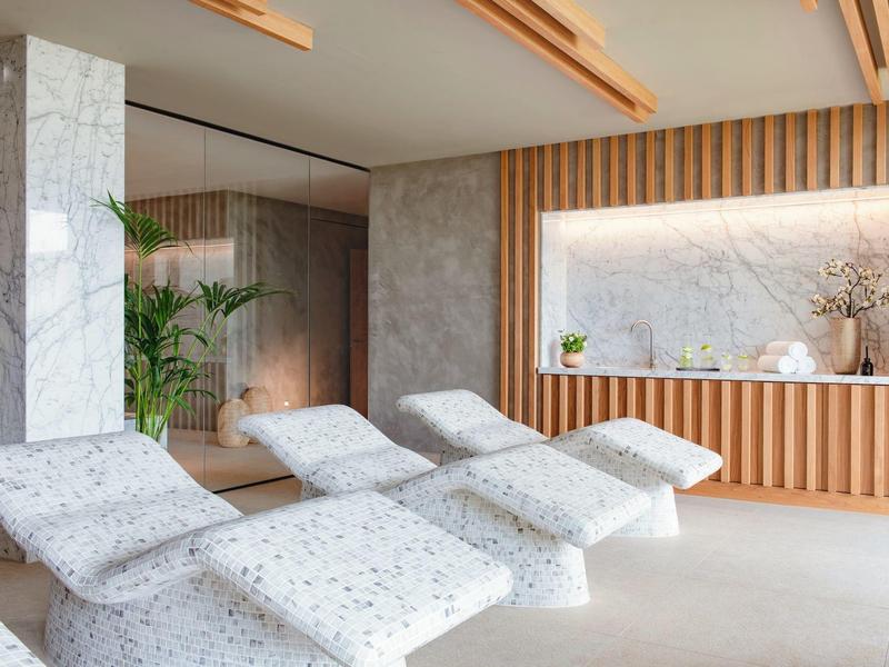 Moderner Ruhebereich mit grauen Liegen, Holzverkleidung und großem Fenster mit Pflanzen und Dekoration.