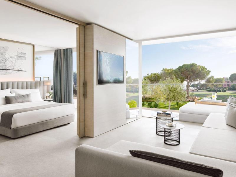 Modernes Hotelzimmer mit Schlafzimmer, Wohnzimmer, weißem Sofa und großem Fenster mit Gartenausblick.