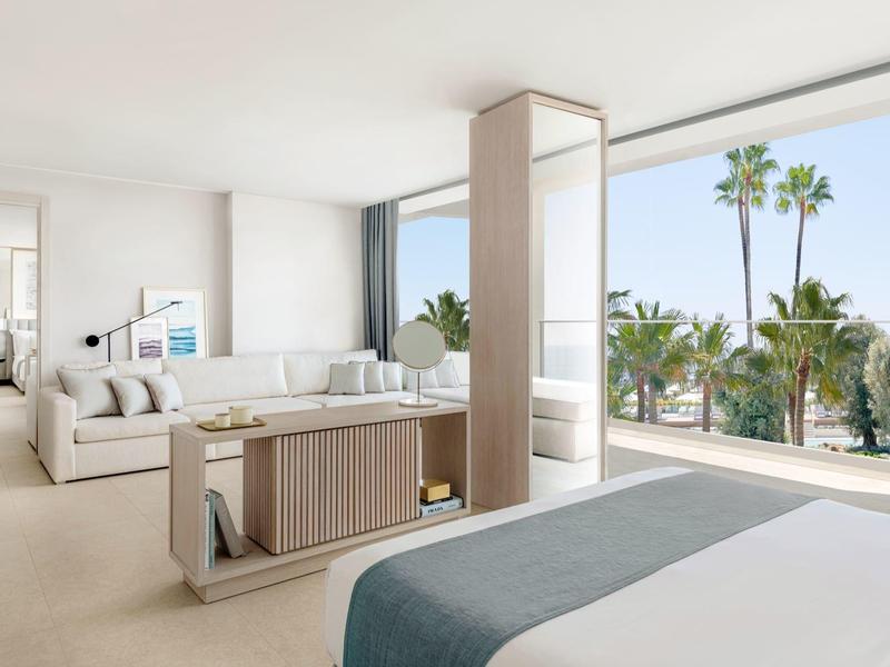 Helles modernes Schlafzimmer mit großem Bett, Sofa, Boden bis zur Decke reichenden Fenstern und Palmenblick.