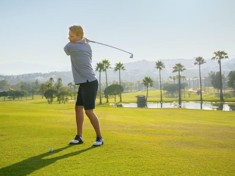 Ein Mann schlägt Golfball auf einem grünen Golfplatz mit Palmen im Hintergrund.