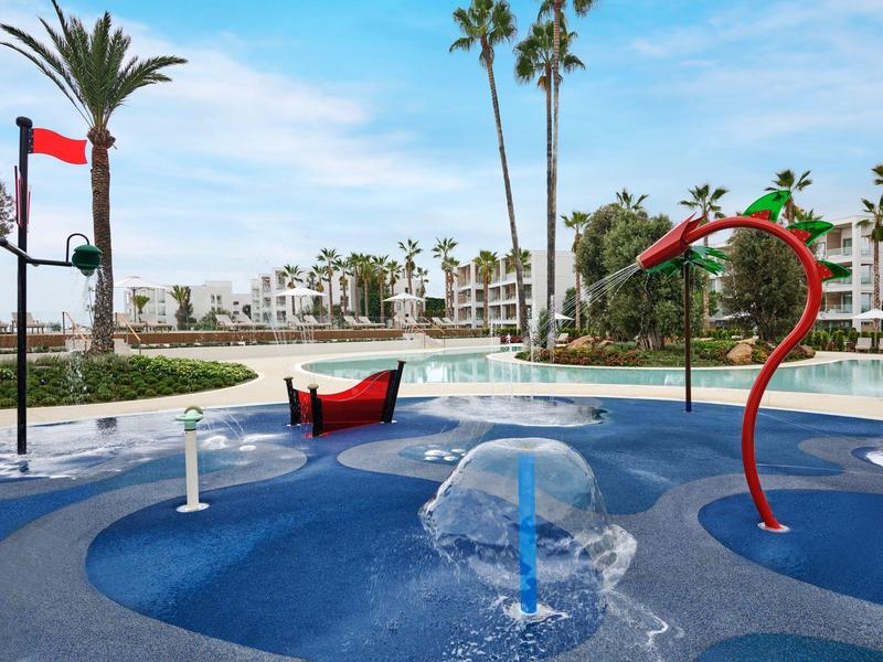 Wasserspielplatz mit bunten Wasserdüsen, Palmen und weißen Gebäuden im Hintergrund.