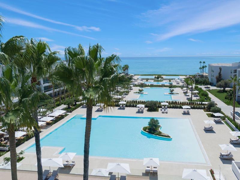 Großer Pool mit palmengesäumter Terrasse und Meerblick unter klarem blauem Himmel.
