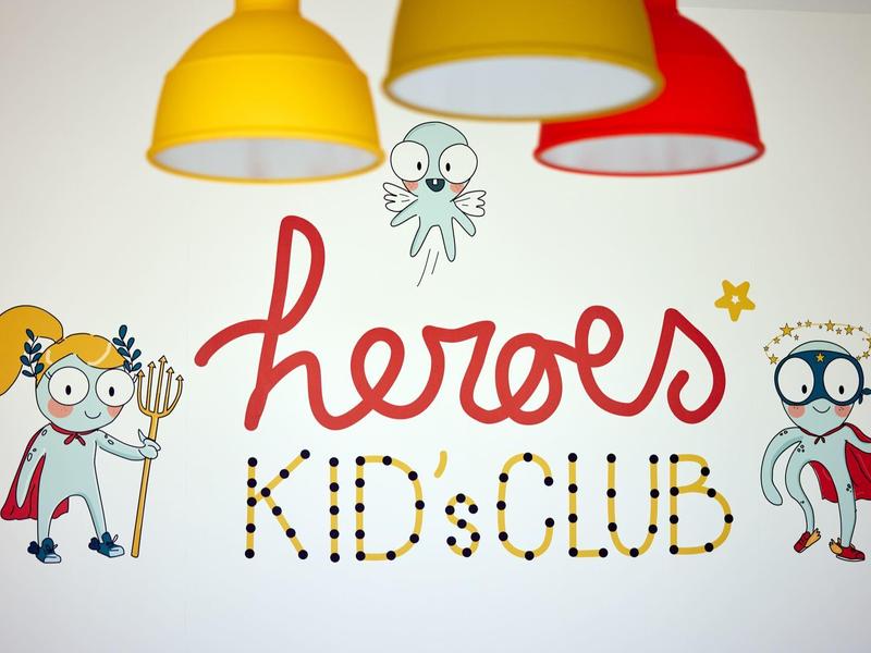 Bunte Wand mit Kinderhelden-Illustrationen und Schriftzug ?heroes KIDS CLUB? unter bunten Lampen.