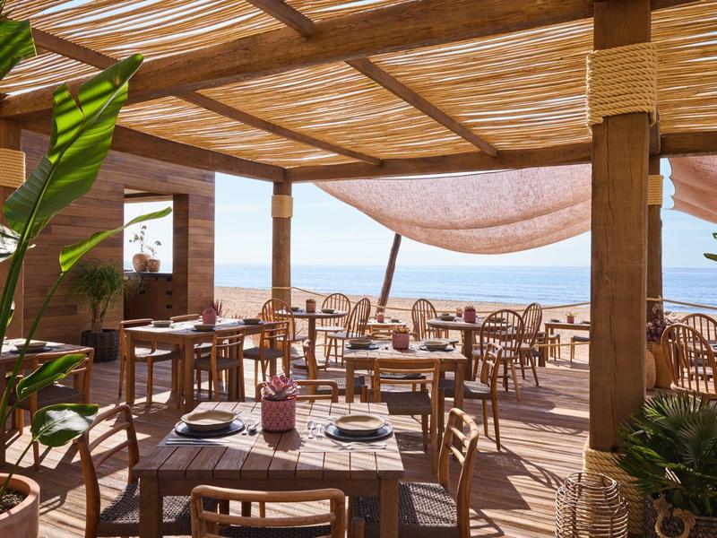 Offene Terrasse mit Holzmöbeln, Pflanzen und Blick auf das blaue Meer unter einem Holzpergola-Dach.