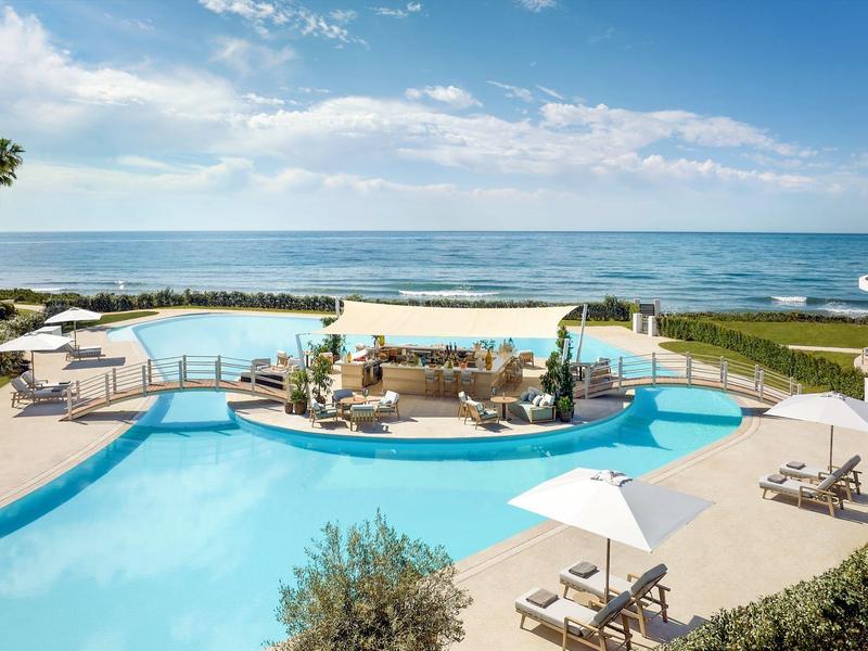 Großer Pool mit Sitzbereich, Liegen, Palmen und Meerblick unter blauem Himmel.