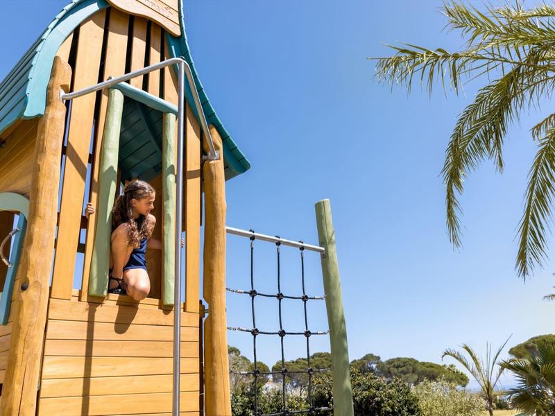 Kind im blauen Badeanzug schaut aus Holzspielhaus bei klarem Himmel und Palmen.