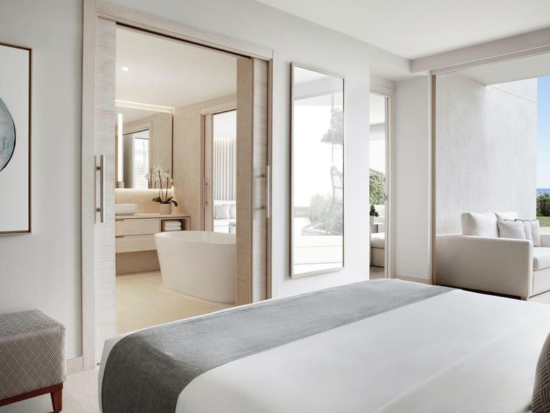 Helles Schlafzimmer mit großem Bett, angrenzendem Badezimmer und Zugang zur Terrasse mit Sitzbereich.