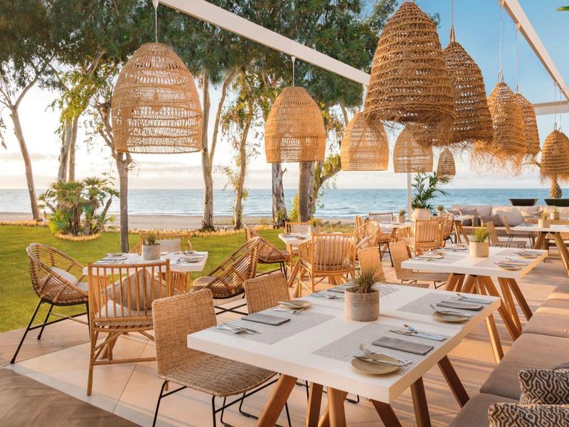 Strandrestaurant mit Holz- und Rattanmöbeln, weißen Tischen und natürlichen Lampenschirmen bei Sonnenuntergang.