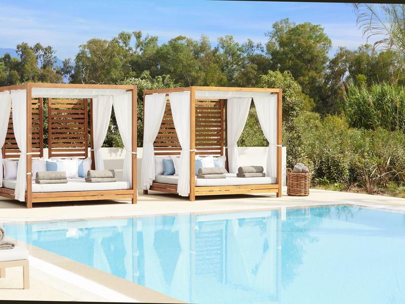 Moderner Pool mit zwei weißen Lounge-Betten, Holzakzenten und grüner Naturkulisse im Hintergrund.