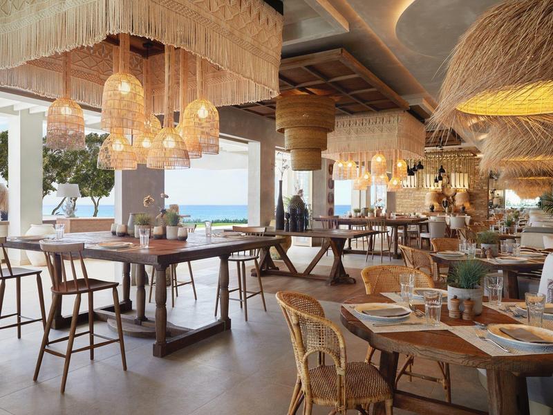 Helles Restaurant mit Holz- und Rattanmöbeln, großen Fenstern und Blick aufs Meer.