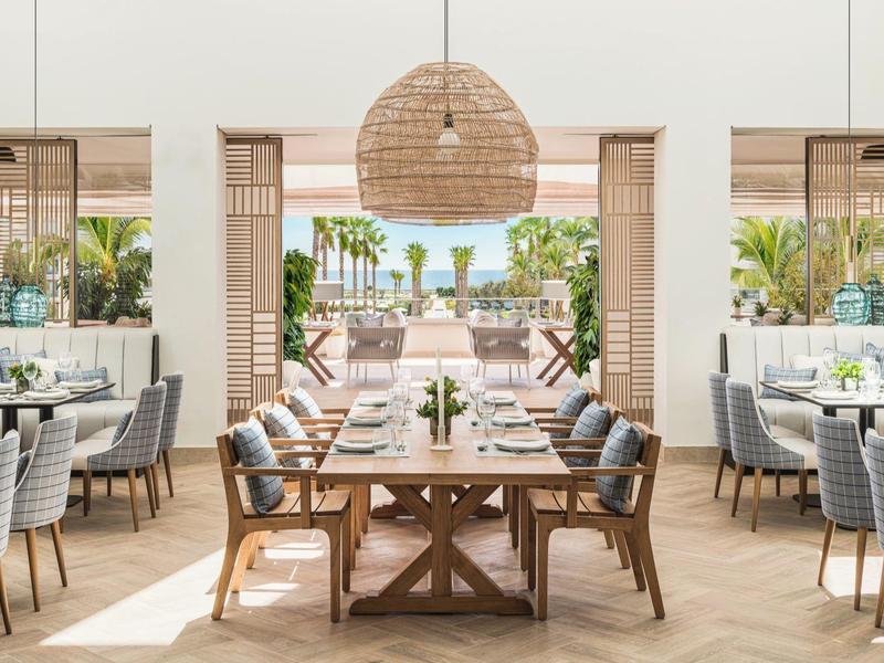 Helles Restaurant mit Holztisch, grauen Stühlen, großen Fenstern und Blick auf Terrasse mit Meer.