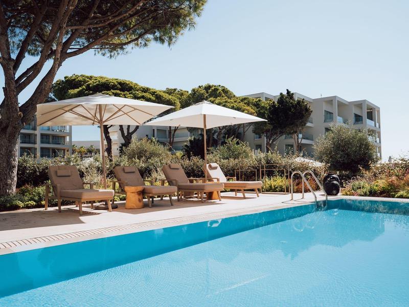 Schwimmbecken mit blauen Wasser, Sonnenschirmen, Liegestühlen und Pflanzen an einem sonnigen Tag.