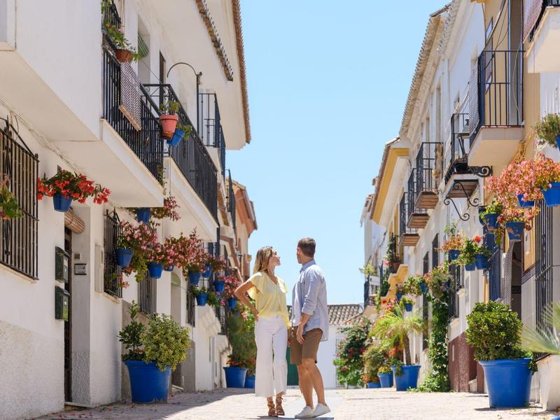 Enges Pflasterstraßendorf mit weißen Häusern, farbigen Blumentöpfen und einem Paar im sommerlichen Outfit.