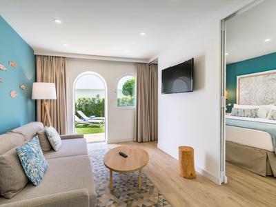 Helles Wohnzimmer mit grauem Sofa, blauem Wandakzent, Holztisch und Blick ins Schlafzimmer und Garten.