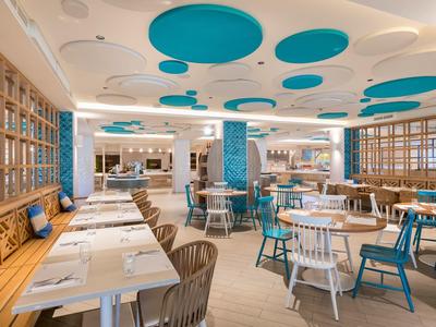 Moderner Gastraum mit weißen Tischen, bunten Stühlen und blauen Deckenelementen in hellem Holzambiente.