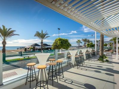 Strandbar mit hohen Tischen und Stühlen, Palmen, Meerblick und sonnigem Himmel.