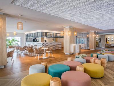 Moderner, heller Raum mit bunten Sitzhockern, Holzboden und großer Fensterfront für viel Tageslicht.