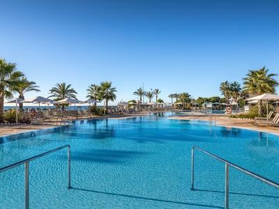 Großer blauer Pool mit Palmen und Sonnenschirmen unter klarem Himmel in tropischer Umgebung.