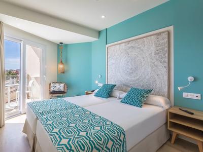 Helles Schlafzimmer mit türkisfarbener Wand, großem Bett, geometrischer Bettdecke und Balkonlicht.