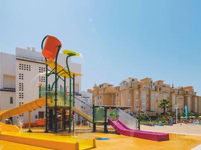 Bunter Wasserspielplatz mit Rutschen und Eimern vor hohen Wohngebäuden unter klarem Himmel.