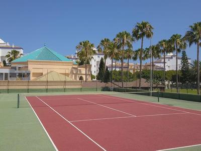 Outdoor tennis court with red surface, surrounded by palm trees and buildings under clear sky.