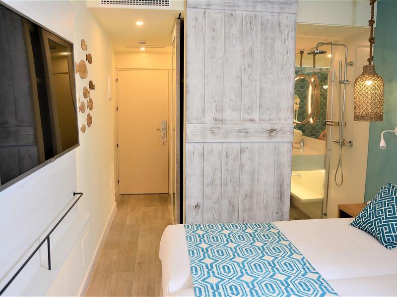 Kleines Schlafzimmer mit Bett, blau gemustertem Bettwäsche-Set, grauem Schrank und offenem Bad mit Spiegel.