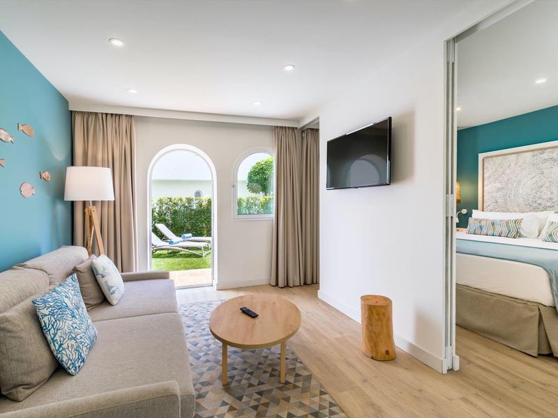 Helles Wohnzimmer mit grauem Sofa, blauem Wandakzent, Holztisch und Blick ins Schlafzimmer und Garten.