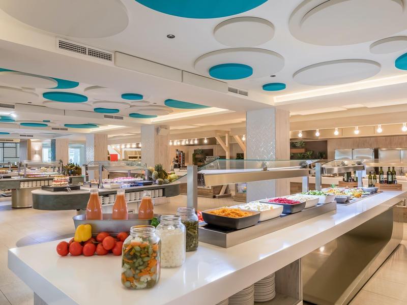 Moderne, helle Kantine mit runden Deckenmustern, langen Buffets und Obst- sowie Snackstationen.