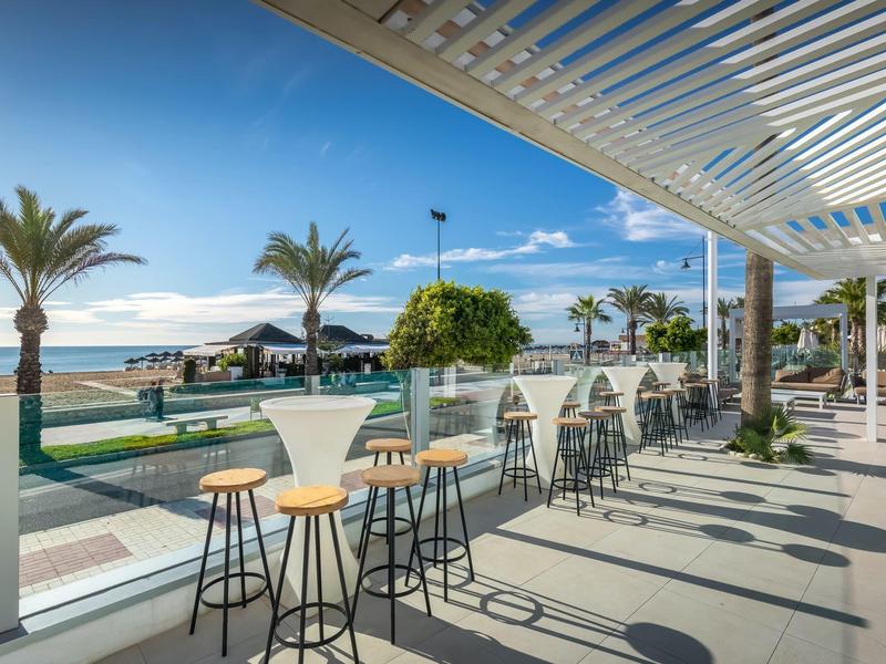 Strandbar mit hohen Tischen und Stühlen, Palmen, Meerblick und sonnigem Himmel.