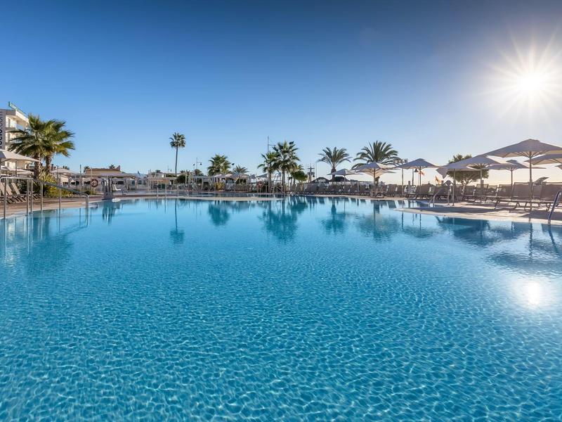 Großer, klarer Pool mit Liegen, Palmen und Sonnenschirmen unter strahlendem blauem Himmel.