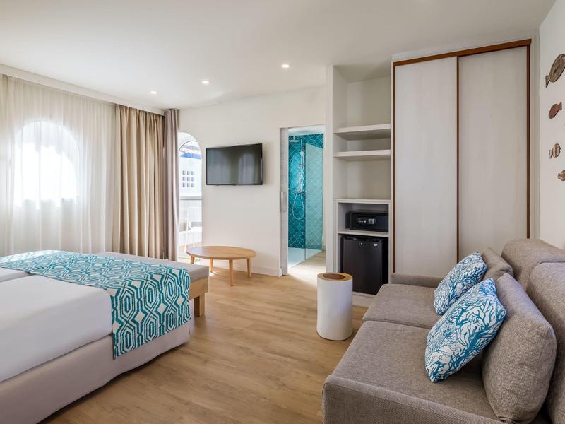 Modern eingerichtetes Hotelzimmer mit Doppelbett, Sofa, Schrank und hellen Holzböden.