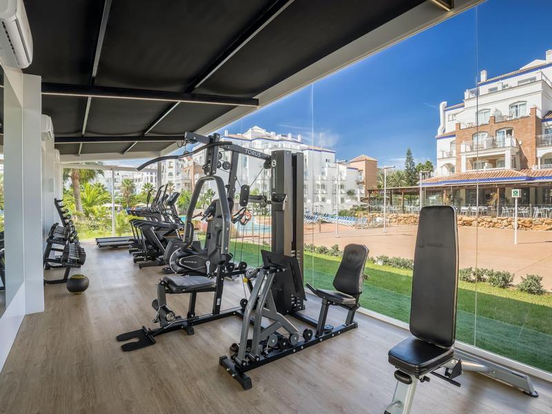 Offenes Fitnessstudio mit Kraftgeräten, großen Fenstern und Blick auf eine grüne Außenanlage.