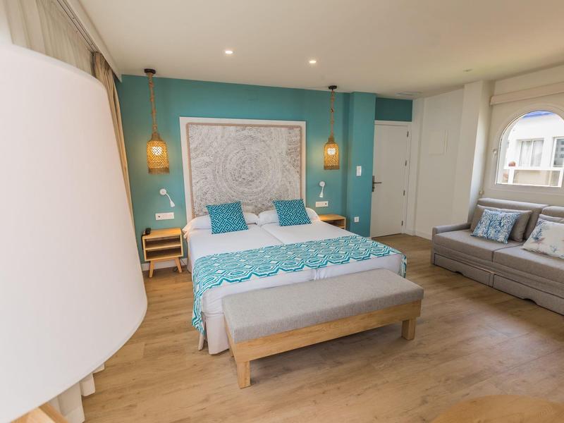 Modernes Schlafzimmer mit Holzboden, türkisem Akzentwand, Doppelbett, Sofa und großen Fenstern.