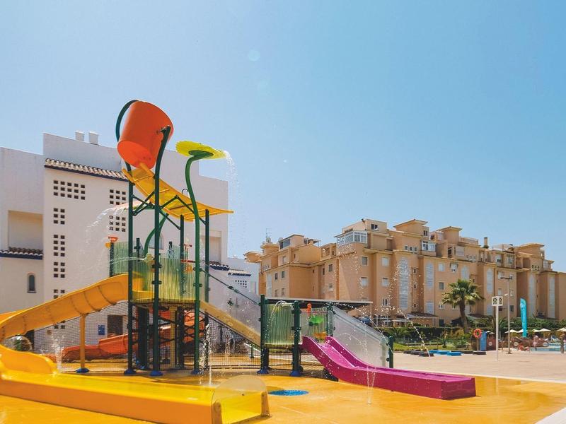 Bunter Wasserspielplatz mit Rutschen und Eimern vor hohen Wohngebäuden unter klarem Himmel.