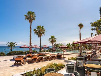 Luxuriöser Hotelpoolbereich mit Sonnenliegen, Palmen und Meerblick unter blauem Himmel.