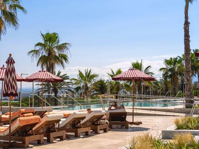 Poolbereich mit Liegestühlen, Sonnenschirmen und Palmen an einem sonnigen Tag.