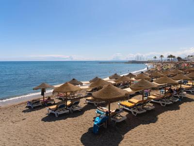 Sandstrand mit Liegestühlen und Strohdächern, ruhiges Meer und klarer blauer Himmel im Hintergrund.