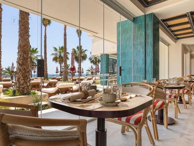 Terrasse eines Cafés mit Tischen und Stühlen unter Glasdach, Palmen im Hintergrund.