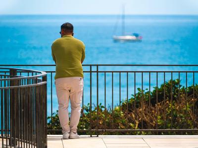 Ein Mann lehnt am Geländer und blickt auf das blaue Meer mit einem Segelboot im Hintergrund.