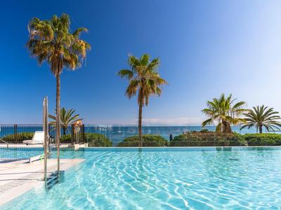 Blauer Swimmingpool mit Palmen, klarer Himmel und Meerblick im Hintergrund.