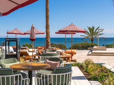 Outdoor-Sitzbereich mit roten Sonnenschirmen, grünen Stühlen und Meerblick bei klarem Himmel.