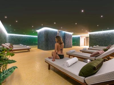 Moderner Ruheraum mit Liegen, grünen Wandflächen, sanfter Beleuchtung und einer sitzenden Frau.