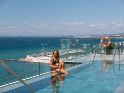 Frau sitzt am Rand eines Infinity-Pools mit Meerblick unter blauem Himmel.