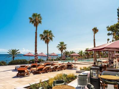 Sonnenterrasse mit Liegestühlen, Palmen, Sonnenschirmen und Blick auf das blaue Meer.