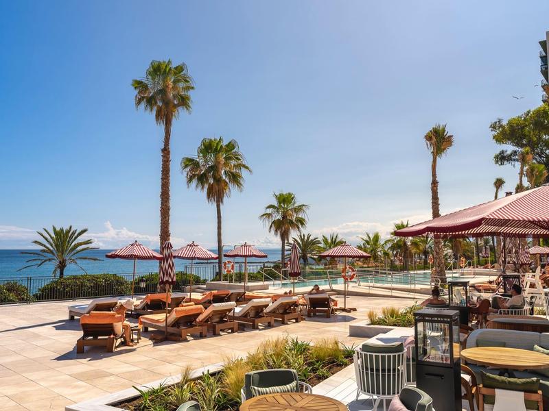 Hotelterrasse mit Sonnenliegen, Palmen, Sonnenschirmen und Meerblick bei klarem Himmel.