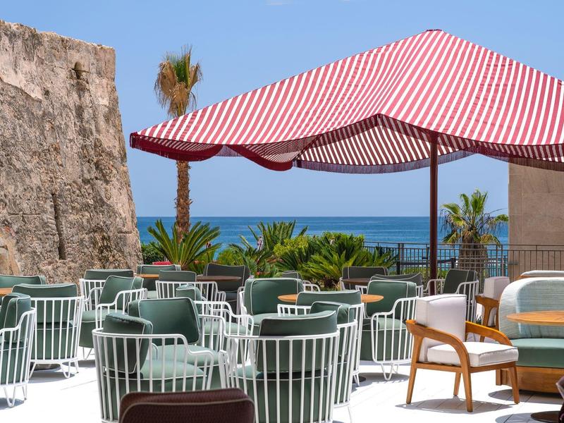 Terrasse mit weißen Metallstühlen, roten Sonnenschirm und Meerblick, Palmen im Hintergrund.
